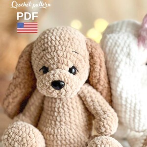 Pu&ograve; includere: Un giocattolo di peluche a forma di cane lavorato a uncinetto in beige e marrone con un naso nero e grandi occhi. Il giocattolo &egrave; seduto su una superficie a strisce bianche e rosa. Schema all'uncinetto PDF.