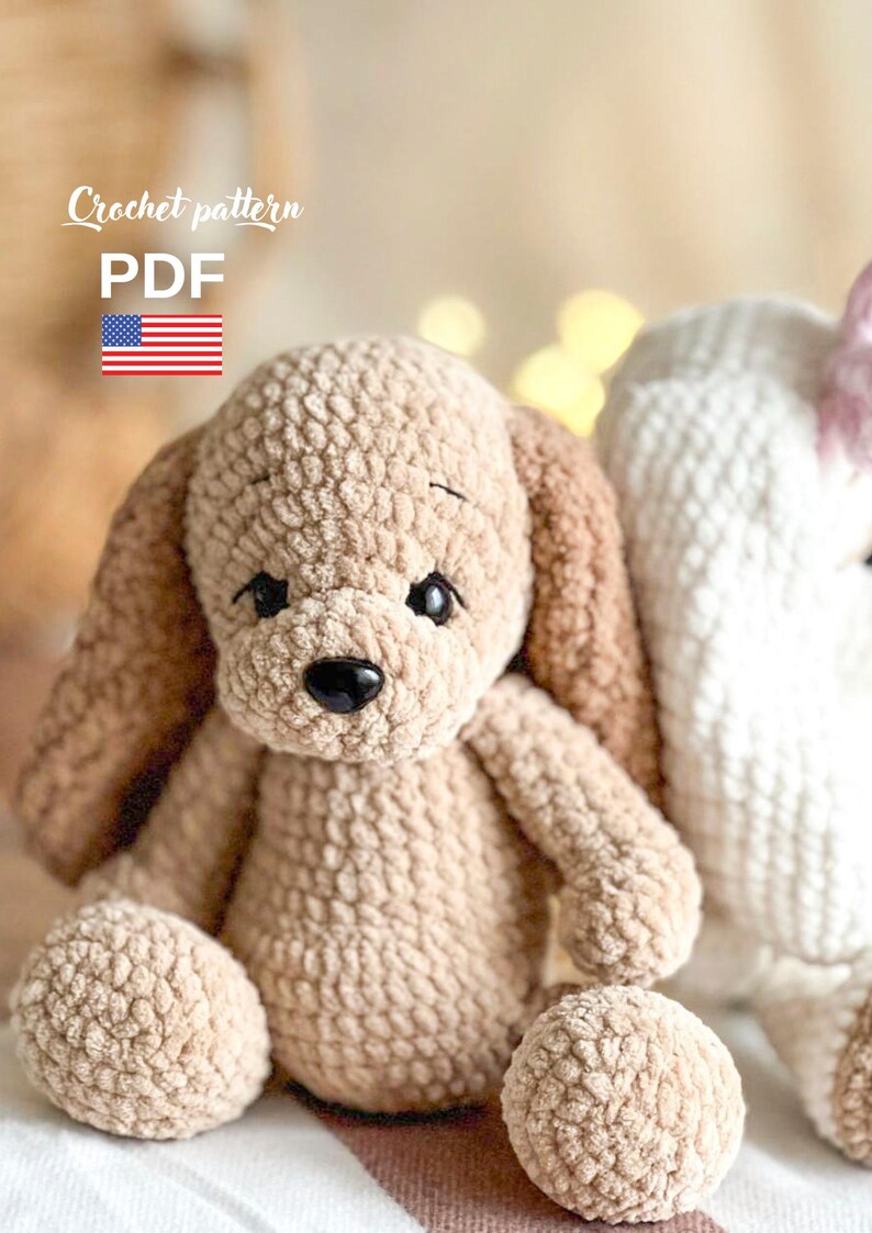 Pu&ograve; includere: Un giocattolo di peluche a forma di cane lavorato a uncinetto in beige e marrone con un naso nero e grandi occhi. Il giocattolo &egrave; seduto su una superficie a strisce bianche e rosa. Schema all'uncinetto PDF.