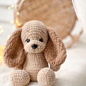 Pu&ograve; includere: Un giocattolo di peluche a forma di cane lavorato a uncinetto con orecchie marroni e corpo beige. Il cane &egrave; seduto su una superficie bianca e soffice.