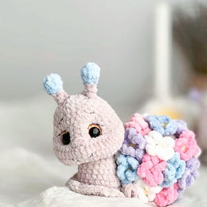 Op de afbeelding: Een gehaakt knuffeldier in de vorm van een slak met een roze lichaam en blauwe antennes. De schelp van de slak is gemaakt van kleurrijke gehaakte bloemen in tinten roze, paars, blauw en wit.