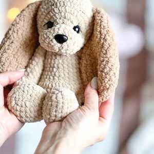 Pu&ograve; includere: Un cane di peluche lavorato a uncinetto con pelliccia marrone e beige. Il cane ha grandi occhi neri e un naso nero.