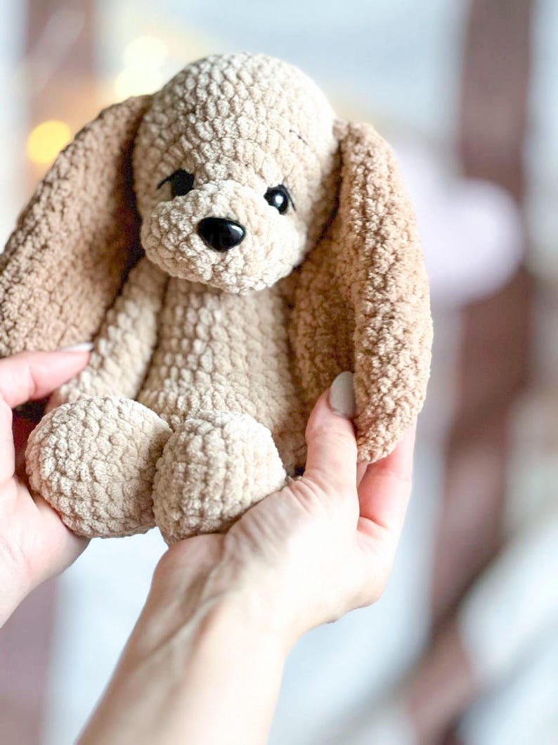 Pu&ograve; includere: Un cane di peluche lavorato a uncinetto con pelliccia marrone e beige. Il cane ha grandi occhi neri e un naso nero.