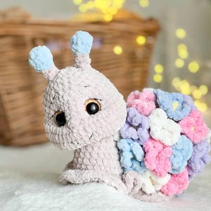 Op de afbeelding: Een gehaakt knuffeldier in de vorm van een slak met een lichtgrijs lichaam, blauwe antennes en een schelp gemaakt van kleurrijke gehaakte bloemen in tinten roze, paars en blauw.