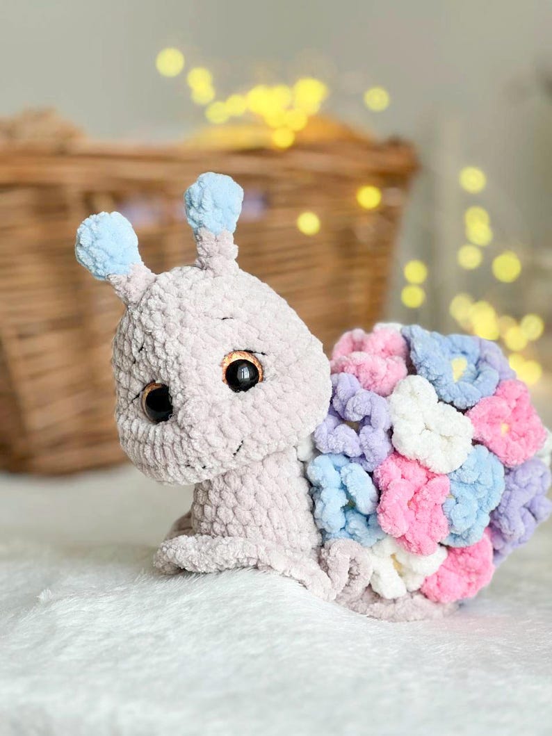 Op de afbeelding: Een gehaakt knuffeldier in de vorm van een slak met een lichtgrijs lichaam, blauwe antennes en een schelp gemaakt van kleurrijke gehaakte bloemen in tinten roze, paars en blauw.