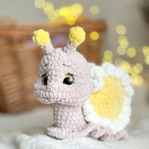Op de afbeelding: Een gehaakt knuffeldier in de vorm van een slak met een roze lichaam, gele antennes en een gele en witte schelp. De slak heeft grote, zwarte ogen met gouden accenten.