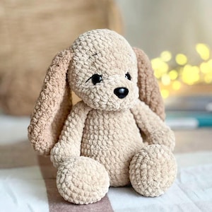 Pu&ograve; includere: Un cane di peluche lavorato a uncinetto con orecchie floppy, occhi marroni e naso nero. Il cane &egrave; seduto su una superficie a strisce bianche e marroni.