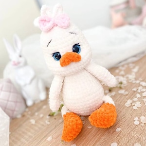 Puede incluir: Un juguete de peluche de pato blanco tejido a crochet con patas naranjas y un lazo rosa. El pato tiene ojos azules y un pico blanco.
