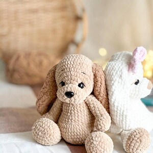 Pu&ograve; includere: Due animali di peluche all'uncinetto, un cucciolo marrone e beige e un agnello bianco, seduti su una coperta a strisce bianche e marroni.