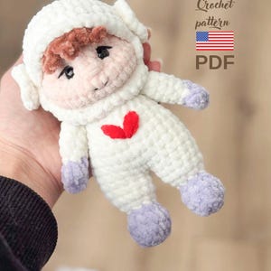 May include: A handmade crochet doll in the shape of a lamb, wearing a white outfit with lavender feet and a red heart. The doll has a white hood and brown hair. Text on the image says "Crochet pattern" and "PDF".