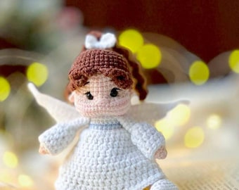 May include: A crocheted angel doll with brown hair, a white dress, and wings. The doll is sitting on a book with a white fuzzy surface in the background.
