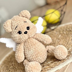 Puede incluir: Un oso de peluche tejido a crochet con alas blancas, sentado en una estera tejida. El oso tiene una textura suave y marrón y ojos negros.