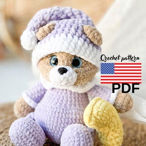 Può includere: Un orsacchiotto all'uncinetto fatto a mano con un completo e un cappello lavanda con finiture bianche, che tiene in mano una banana gialla all'uncinetto. L'orso ha gli occhi blu. L'immagine include il testo "Crochet pattern" e "PDF".