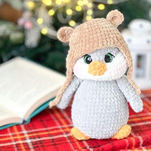 Può includere: Una bambola pinguino all'uncinetto fatta a mano con un cappello da orso marrone. Il pinguino ha una faccia bianca, corpo grigio, becco e piedi arancioni e occhi verdi. È posizionato su una superficie a quadri rossi.