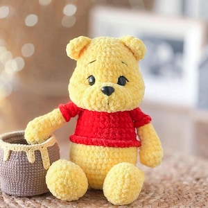 May include: A crocheted Winnie the Pooh plush toy wearing a red shirt. The toy is sitting on a woven mat next to a brown crocheted honey pot.