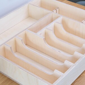 May include: A light-colored wooden drawer organizer with multiple compartments. The organizer has curved dividers and rectangular sections, designed to hold various items. The wood grain is visible, and the construction appears sturdy.