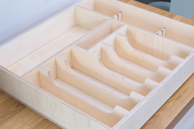 May include: A light-colored wooden drawer organizer with multiple compartments. The organizer has curved dividers and rectangular sections, designed to hold various items. The wood grain is visible, and the construction appears sturdy.