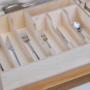 May include: A wooden cutlery organizer with multiple compartments, holding various silverware pieces. The silverware includes spoons, forks, and knives. The organizer is made of light-colored wood and sits on a wooden surface.