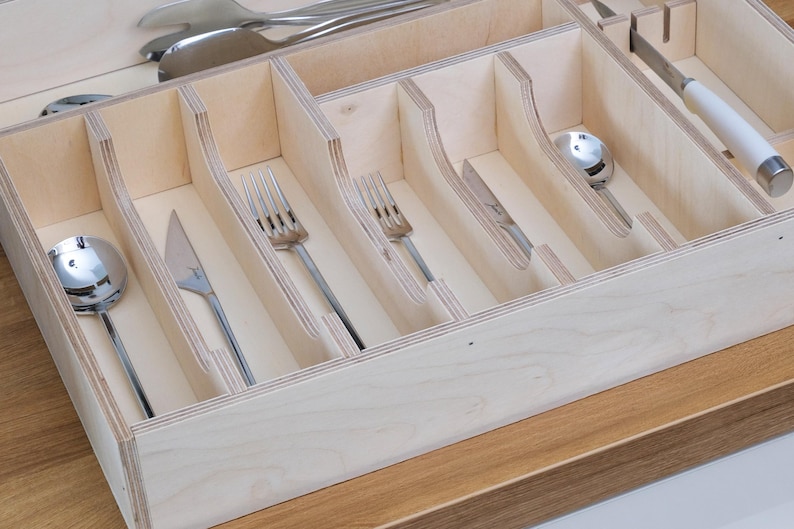 May include: A wooden cutlery organizer with multiple compartments, holding various silverware pieces. The silverware includes spoons, forks, and knives. The organizer is made of light-colored wood and sits on a wooden surface.