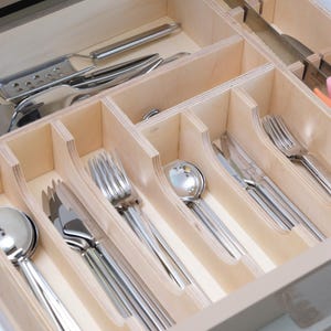 May include: A wooden drawer organizer filled with stainless steel silverware. The drawer contains spoons, forks, knives, and serving utensils. Some knives have white, pink, and orange handles. The organizer is made of light-colored wood.