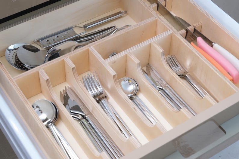 May include: A wooden drawer organizer filled with stainless steel silverware. The drawer contains spoons, forks, knives, and serving utensils. Some knives have white, pink, and orange handles. The organizer is made of light-colored wood.