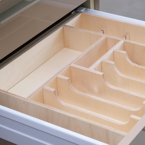 May include: Open wooden drawer with a custom-built organizer. The organizer features multiple compartments of varying sizes, including curved slots, designed for cutlery or kitchen utensils. The drawer and organizer are made of light-colored wood.
