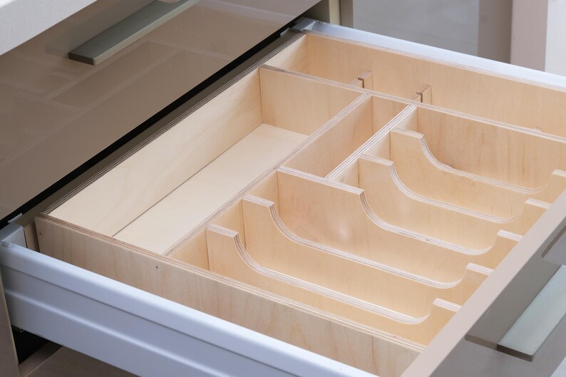 May include: Open wooden drawer with a custom-built organizer. The organizer features multiple compartments of varying sizes, including curved slots, designed for cutlery or kitchen utensils. The drawer and organizer are made of light-colored wood.