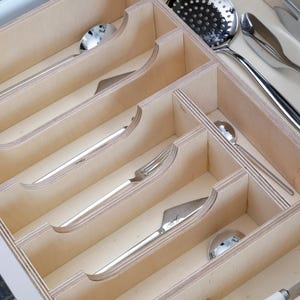 May include: A wooden cutlery organizer with multiple compartments, holding various stainless steel utensils. The silverware includes spoons, forks, knives, and a slotted spoon. The organizer is made of light-colored wood.