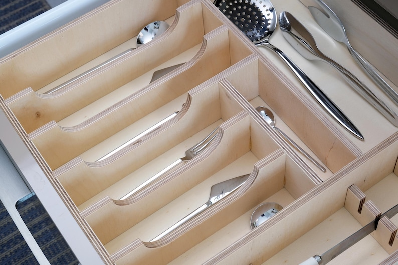 May include: A wooden cutlery organizer with multiple compartments, holding various stainless steel utensils. The silverware includes spoons, forks, knives, and a slotted spoon. The organizer is made of light-colored wood.