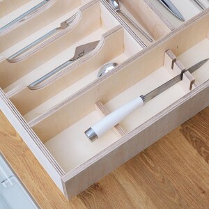 May include: Wooden cutlery organizer with multiple compartments. The organizer holds various silverware pieces, including forks, knives, and spoons. The knife has a white handle and a silver blade. The organizer is made of light-colored wood.