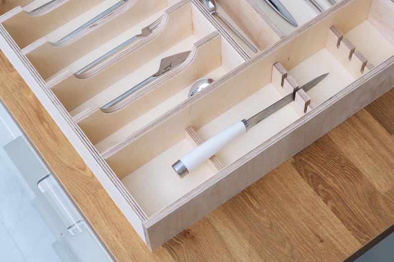 May include: Wooden cutlery organizer with multiple compartments. The organizer holds various silverware pieces, including forks, knives, and spoons. The knife has a white handle and a silver blade. The organizer is made of light-colored wood.