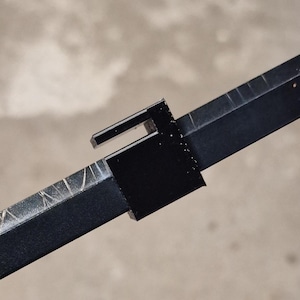 Pode incluir: Close-up de um objeto de metal preto com formato retangular e uma extensão em forma de gancho. O objeto tem um acabamento fosco e parece ser um componente de uma estrutura maior. O fundo é um tom neutro desfocado.