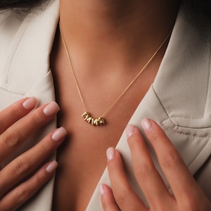 Puede incluir: Un collar de oro con un colgante de placa con el nombre "AMMA". El collar lo lleva una persona con una chaqueta marrón claro.