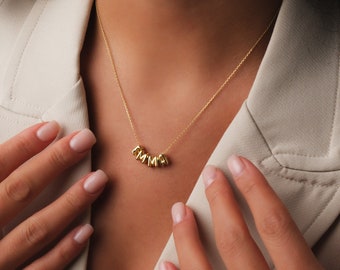 May include: A gold necklace with a nameplate pendant spelling out "AMMA". The necklace is worn by a person wearing a light brown blazer.
