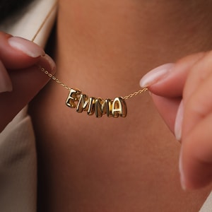 Könnte beinhalten: Eine goldene Halskette mit dem Namen "EMMA", der in einzelnen Buchstaben geschrieben ist.