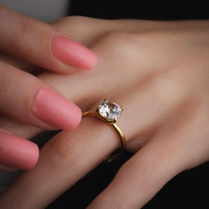May include: A gold ring with a large, clear, round-cut diamond. The ring is on a finger with pink nail polish.