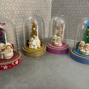 May include: Four glass dome ornaments with a Christmas nativity scene inside. Each dome has a different coloured base and a small Christmas tree inside. The trees are decorated with red and white baubles, and the nativity scenes feature a white baby Jesus, Mary, and Joseph.