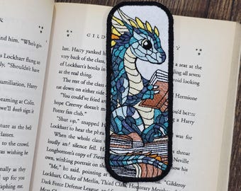May include: Embroidered bookmark featuring a blue and teal dragon reading a book. The dragon has yellow accents and is set against a white background with a black border. The bookmark is placed in an open book.