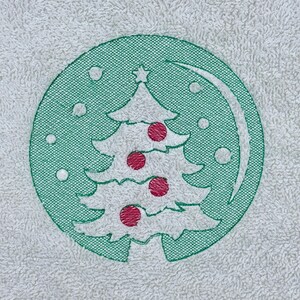Könnte beinhalten: Weißes Handtuch mit einem gestickten Weihnachtsbaum-Design. Der Baum ist weiß mit roten Kugeln und einem Stern, eingefasst in einem grünen Kreis. Kleine weiße Punkte umgeben den Baum und schaffen ein festliches Urlaubsgefühl.