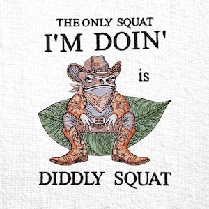 Könnte beinhalten: Gesticktes Design mit einem Frosch in Cowboyhut und -stiefeln, der auf einem Blatt sitzt. Der Text lautet "THE ONLY SQUAT I'M DOIN' is DIDDLY SQUAT". Der Frosch trägt ein Halstuch und einen Gürtel. Das Design hat einen weißen Hintergrund.