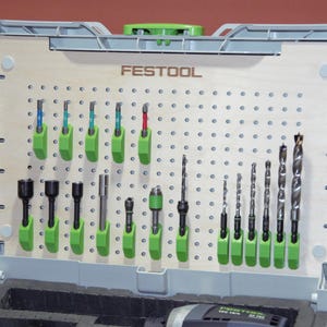 Op de afbeelding: Een lichtgrijze Festool opbergkoffer met een lichthouten pegboard-inzetstuk met het Festool-logo. Het pegboard bevat diverse metalen boortjes en schroefbits, vastgezet door felgroene kunststof houders die in rijen zijn geplaatst.