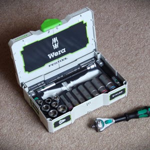 Puede incluir: Un juego de herramientas Wera abierto con varios vasos y una llave de carraca. La caja es blanca y gris con detalles verdes y el logotipo de Wera. El juego incluye múltiples vasos y una llave de carraca.