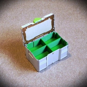 Op de afbeelding: Een open grijze en groene plastic opbergdoos met het woord "Festool" gedrukt op de deksel. De doos heeft vijf compartimenten met groene scheidingswanden.