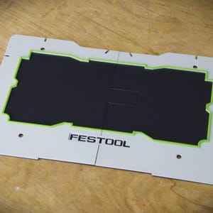 Könnte beinhalten: Weiße Kunststoffschablone mit grünem Umriss und dem aufgedruckten Wort "FESTOOL". Die Schablone ist zum Ausschneiden einer rechteckigen Form gedacht.