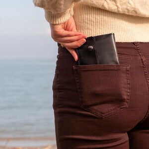 May include: Black leather wallet with a snap closure partially visible in the back pocket of brown pants. The wallet is rectangular and appears to be folded. The person is wearing a cream-colored sweater. The background is a blurred view of the ocean.