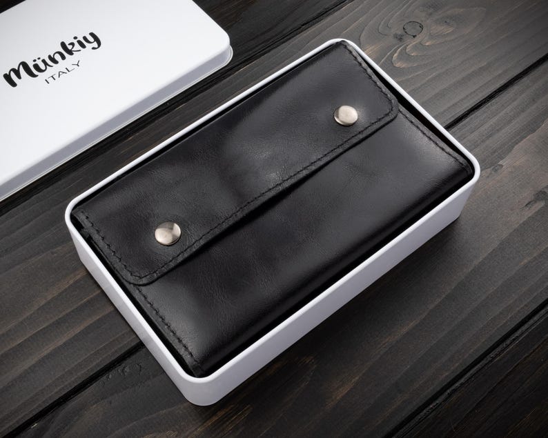 May include: A black leather wallet with a snap closure, presented in a white box. The wallet is rectangular and has a smooth texture. The box has the brand name "M&uuml;nkiy ITALY" printed on the lid.