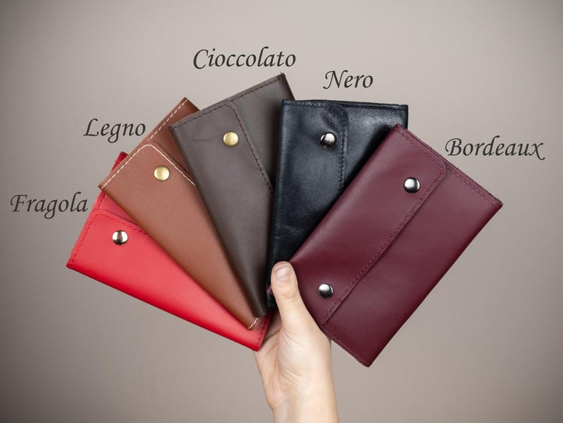 May include: A hand holding a fan of leather wallets in various colors: red, brown, dark brown, black, and burgundy. Each wallet has a snap closure. The wallets are labeled with the Italian words "Fragola", "Legno", "Cioccolato", "Nero", and "Bordeaux".