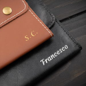 May include: Close-up of two leather cases. One is brown with the initials "S.C." in gold and a gold button closure. The other is black with the name "Francesco" in white script. Both cases are stitched with white thread.
