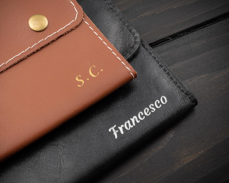 May include: Close-up of two leather cases. One is brown with the initials "S.C." in gold and a gold button closure. The other is black with the name "Francesco" in white script. Both cases are stitched with white thread.