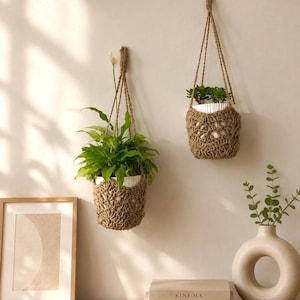 Op de afbeelding: Twee bruine gehaakte plantenhangers van dik touw, elk met een witte pot met een groene plant. De hangers zijn aan de muur bevestigd en hebben een opengewerkt patroon en lange gevlochten koorden om ze op te hangen.