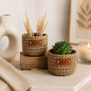 May include: Two round, hand-crocheted baskets made from tan jute twine. Each basket features a small, rectangular brown leather tag embossed with the text snolk. One basket holds dried wheat stalks, while the other contains a green succulent plant.